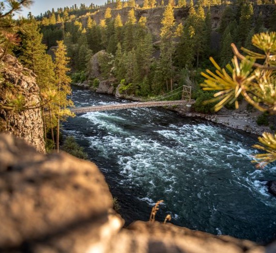 A view from a hiking trail at Bowl & Pitcher Park in Spokane. The view is of the river with a wooden bridge over it and various greenery in the background and rocks in the foreground.