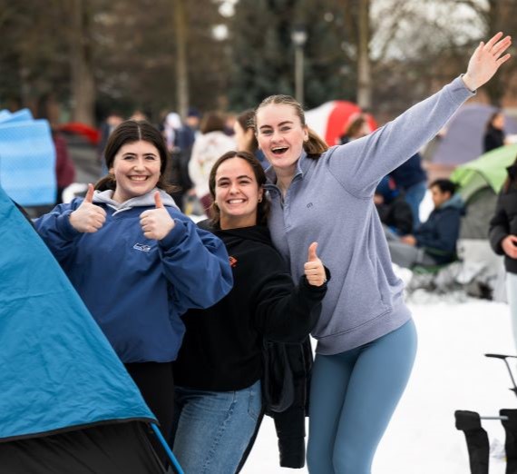 Three young women in college standing smiling for a picture in a sea of tents with snow on the ground at Gonzaga's Kennel Campout tradition.