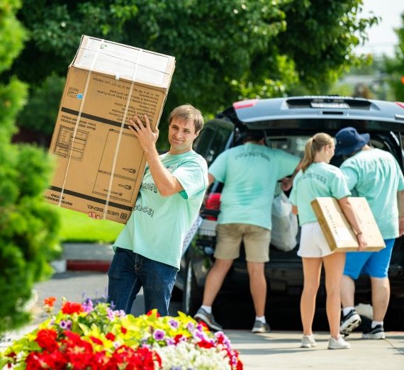 Gonzaga staff member wearing a light green volunteer shirt carrying a large box during move-in weekend. In the background, there are three more volunteers helping take boxes out of the trunk of a car. In the foreground, there is a tree and brightly colored flowers.