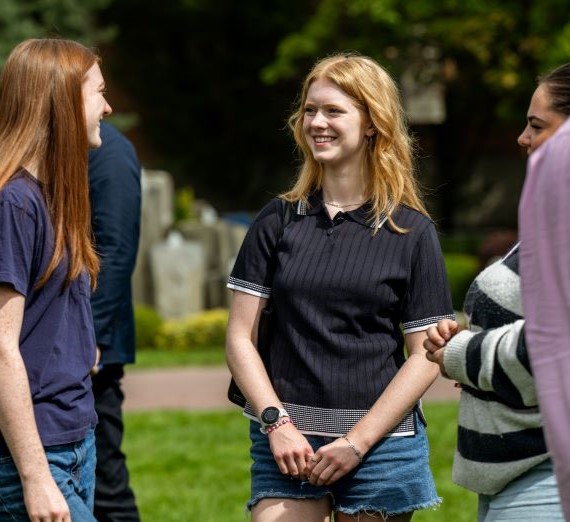 Three students chatting during Welcome Weekend outside. The student in the middle is wearing a black shirt with denim shorts and is smiling chatting with other students.