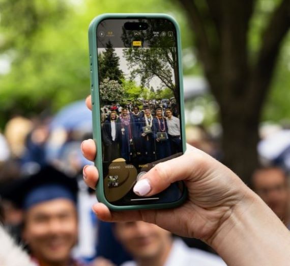 A close-up of someone taking a picture on an iPhone of students standing in blue caps and gowns at a Gonzaga University graduation. The hand holding the phone has pink nail polish on it.