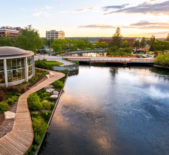 An aerial drone shot of Riverfront Park with the Spokane River flowing, a bridge in the distance, and on the left-hand side, the Looff Carousel and wooden pathway along the river.