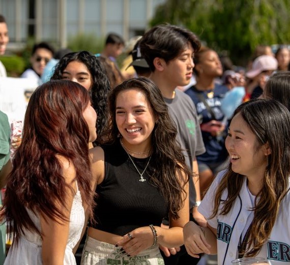 Three students smiling at each other while attending the Gonzaga University Club Fair. They are three female students who are laughing to each other with a crowd of people around them also at the club fair.