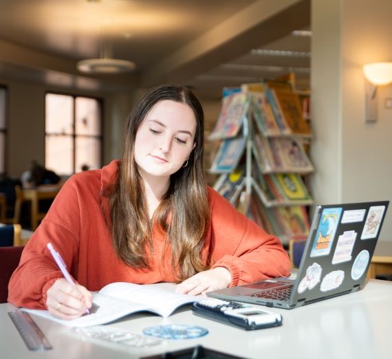 Student wearing an orange sweater writing in her notebook in the Foley Library with her laptop open nearby. The student has long, straight brown hair and is slightly smiling.
