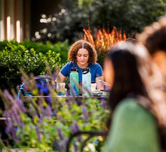A girl studying outside at a lunch table on Gonzaga's campus. She is wearing a blue shirt and has curly hair. There are purple flowers in the foreground.