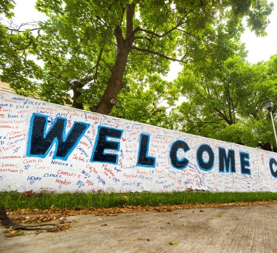 Portion of a wall here on campus with the spray painted word "Welcome" in blue with lots of small written names around it.