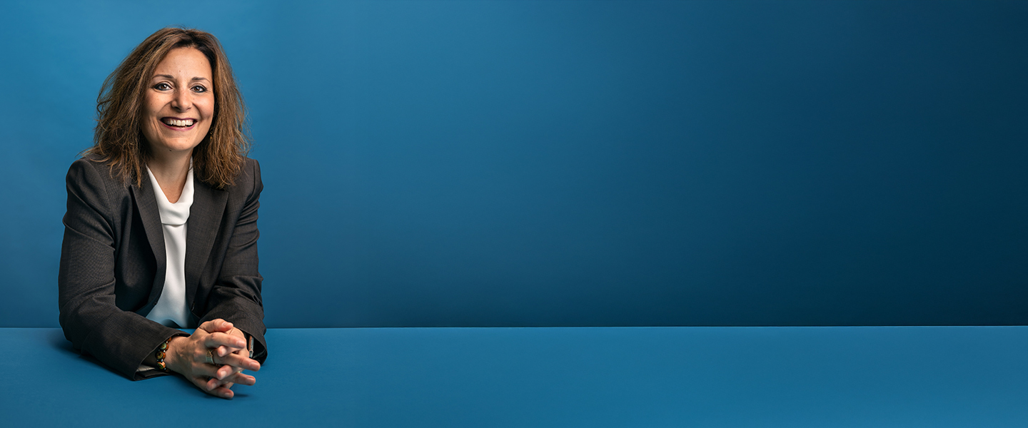  A smiling person, wearing a dark suit jacket and white top, leans on a blue surface against a matching blue background.