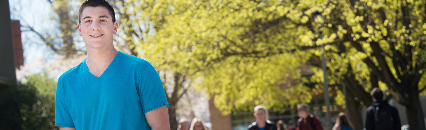 A smiling person in a blue shirt stands outdoors, with other people in the background under bright green trees.